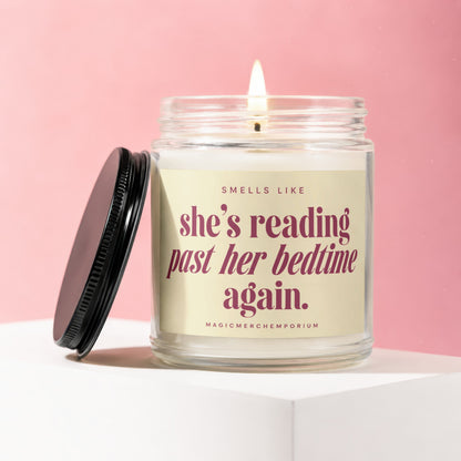 Glass candle with lit wick and black lid, says "SMELLS LIKE she's reading past her bedtime again. MAGICMERCH..."