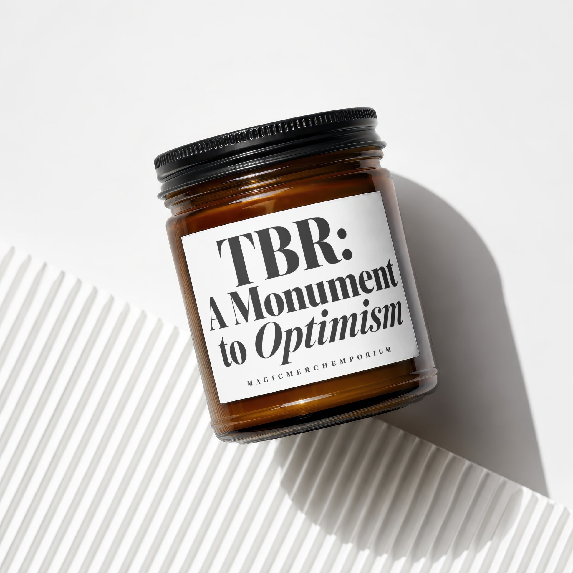 Glass jar candle with black lid features a label reading "TBR: A Monument to Optimism."