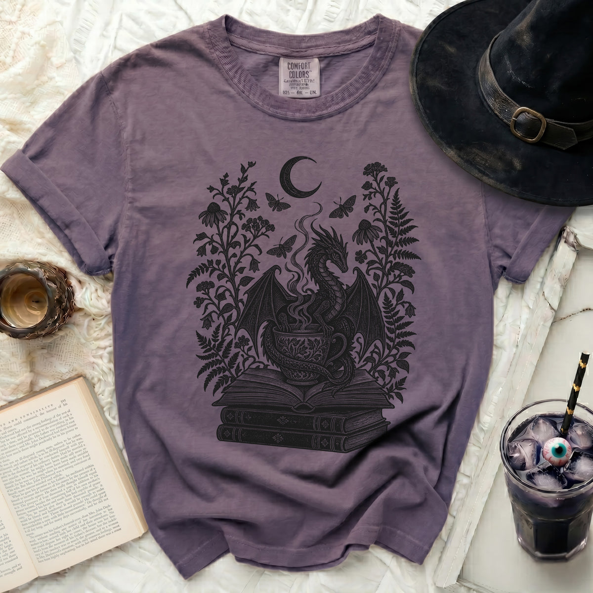 Muted purple Comfort Colors t-shirt with a black line art graphic of a dragon on a stack of books surrounded by floral arrangements, a moon, and moths.