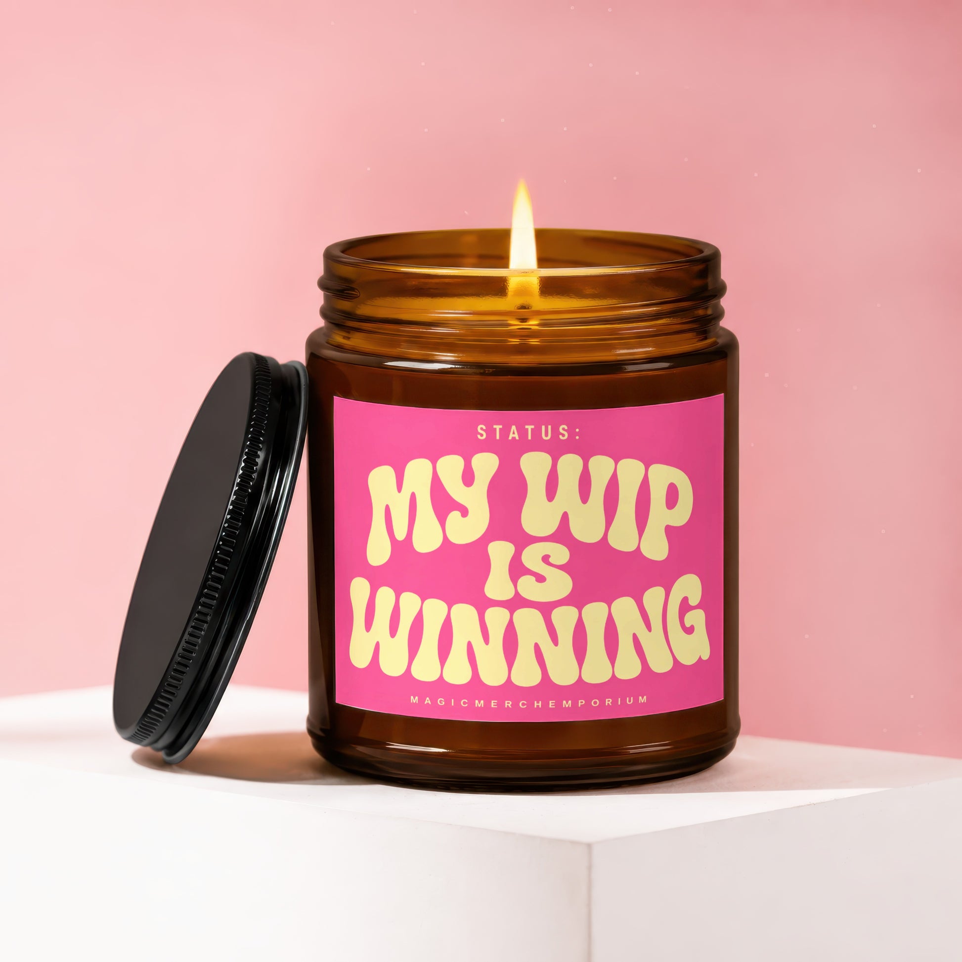 Amber jar candle with lit flame and pink label saying "Status: My WIP is Winning".