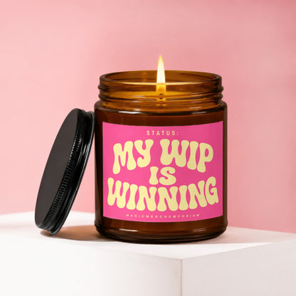 Amber jar candle with lit flame and pink label saying "Status: My WIP is Winning".