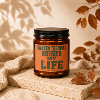 Amber candle with black lid, orange label text: "Cracked the spine, ruined my life." MagicMerchEmporium.
