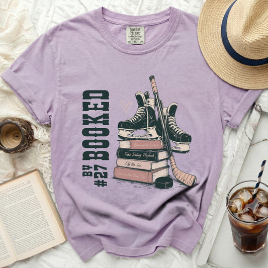 Lavender graphic tee with hockey skates, stick, and books, "Booked" text.