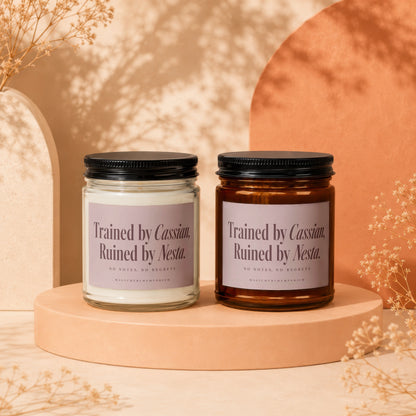 Two scented candles with purple labels reading "Trained by Cassian, Ruined by Nesta" on a beige pedestal.