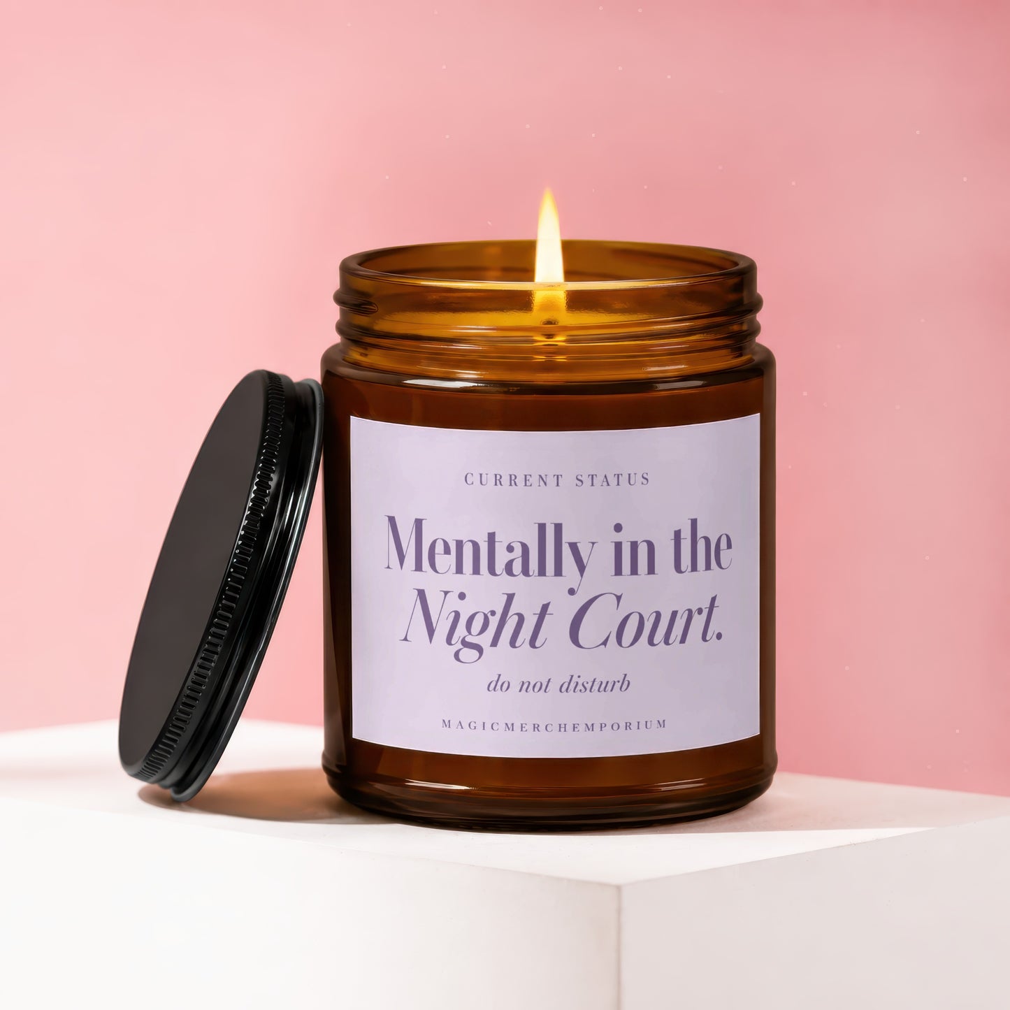 A lit amber glass jar candle with a black lid and white label against a pink background.