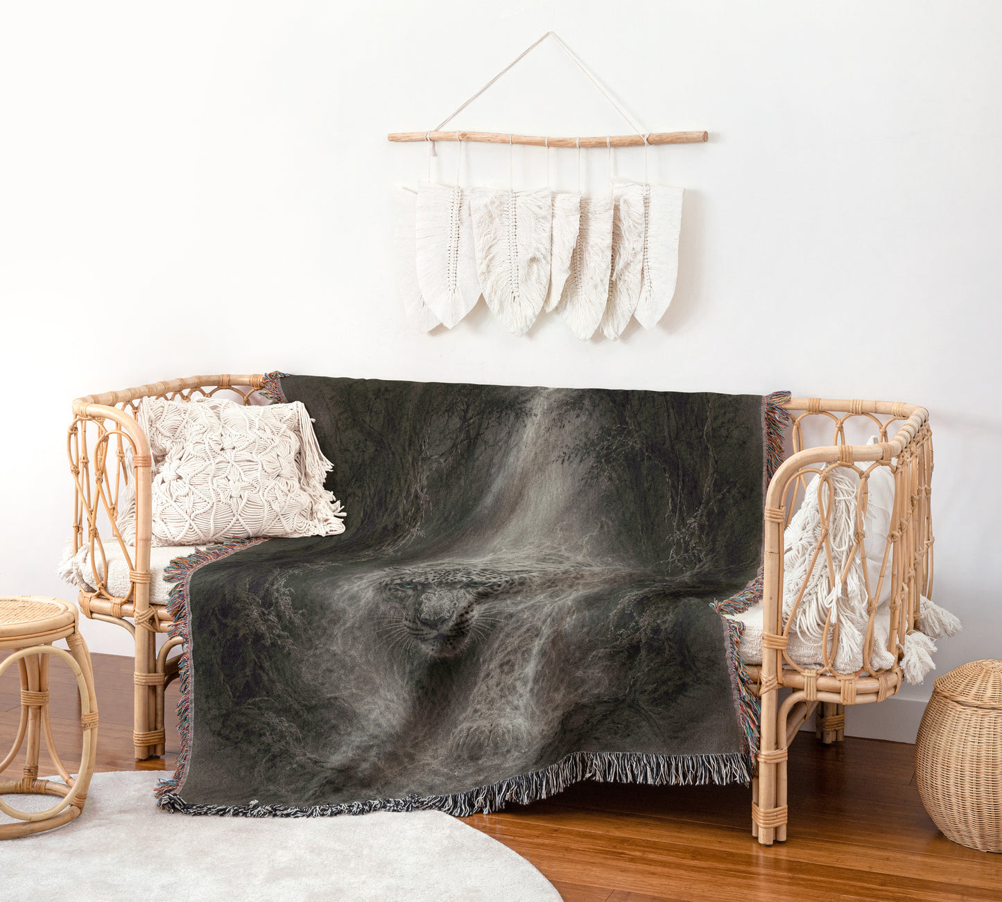 a cozy living room with a wicker sofa adorned with a black and gray throw blanket, a white pillow, and a woven basket. Above the sofa, a wall decoration with white feathers hangs, and a wooden stool is placed nearby.