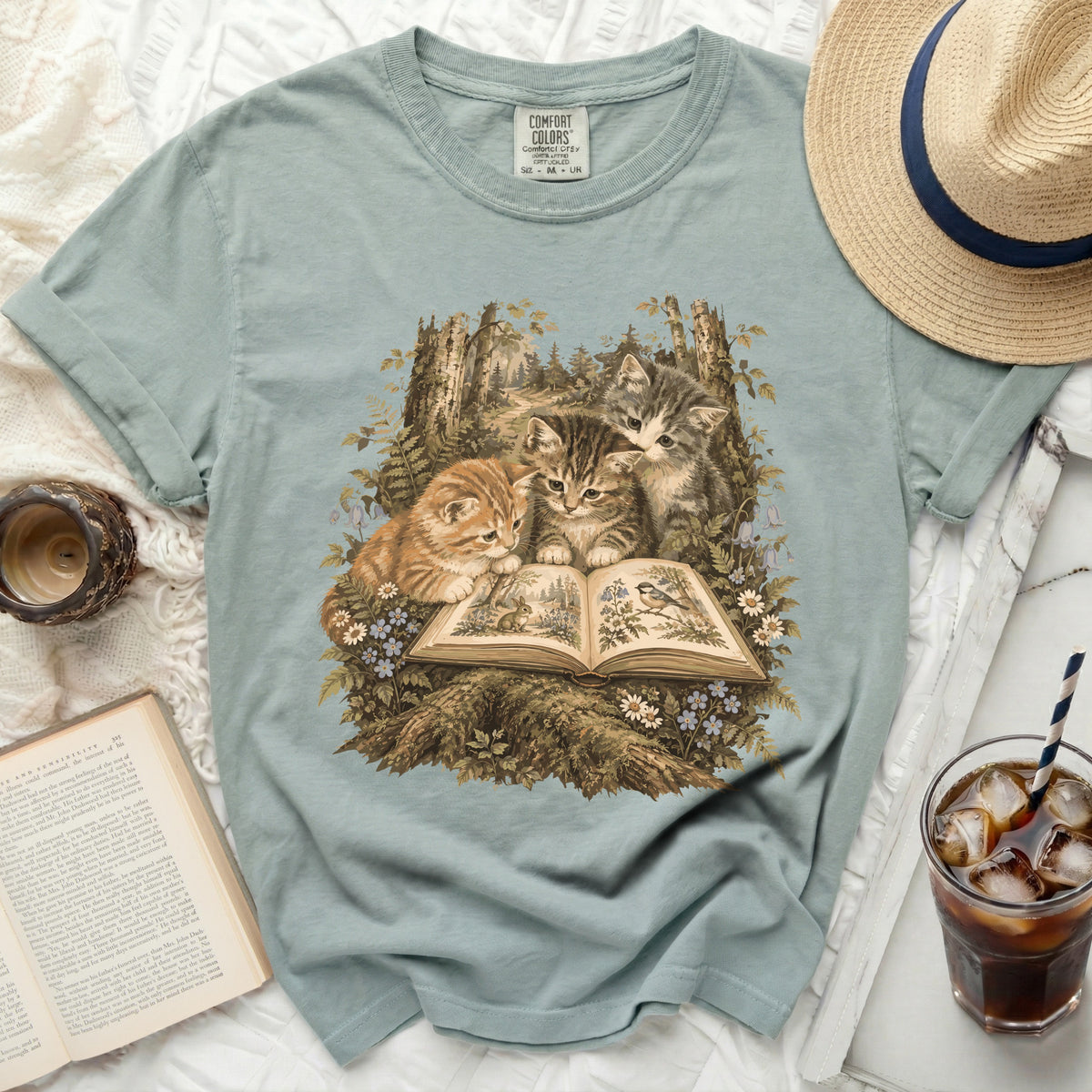 Three kittens look at an open book amidst a forest scene with trees and flowers.