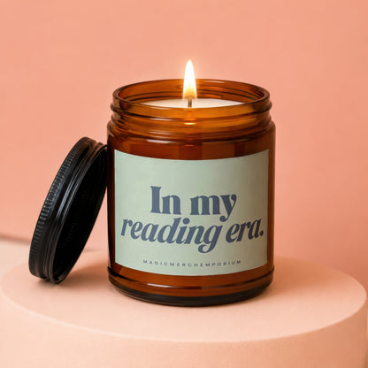 Amber glass candle with lit wick and lid askew, reads "In my reading era."