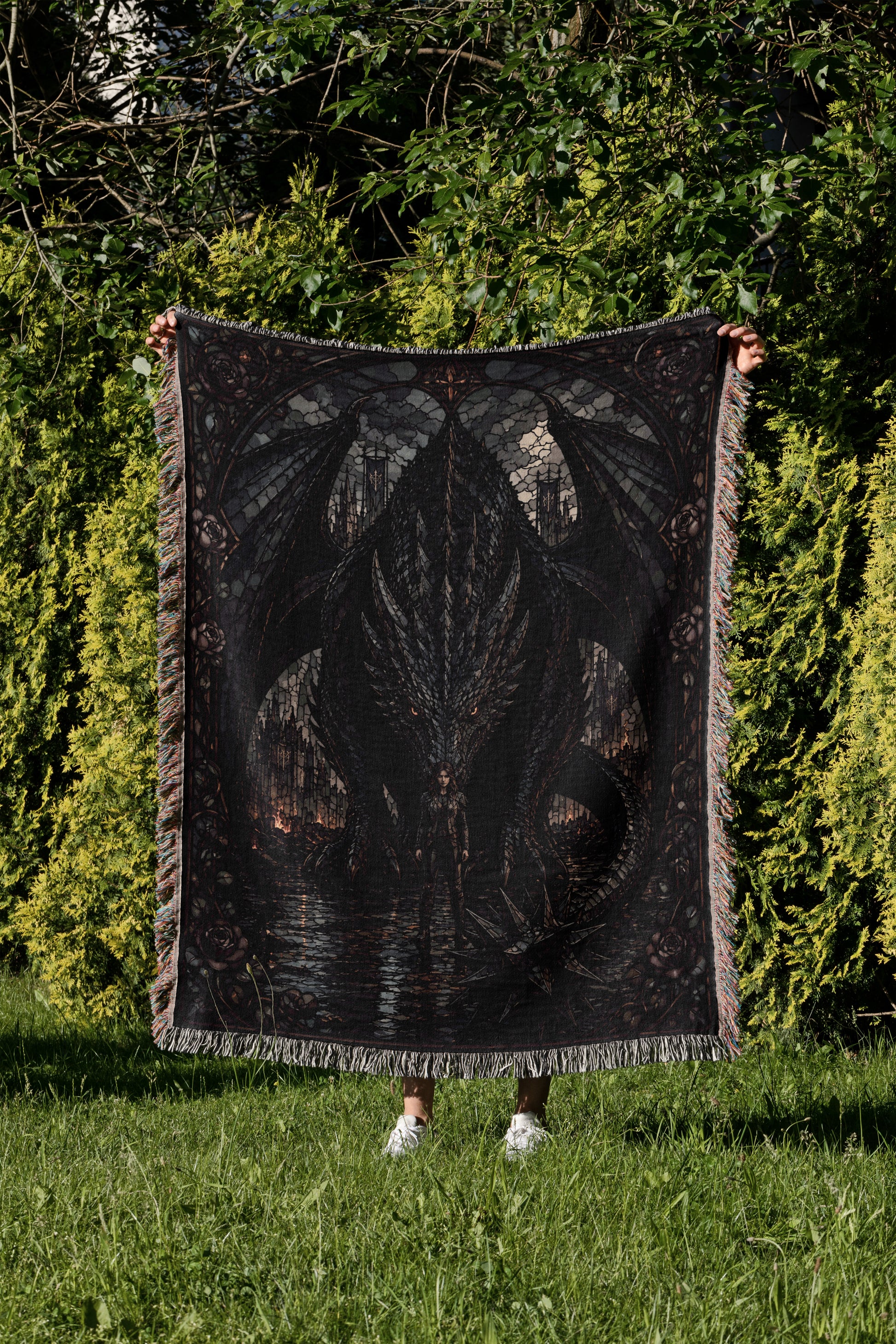 A person is standing in a grassy area, holding up a large, intricately designed blanket featuring a dragon design.