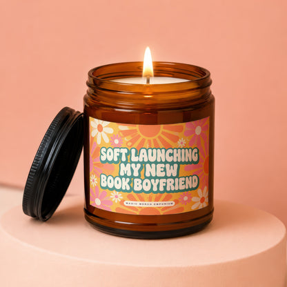 Amber candle with a lit wick and a black lid, featuring a label that reads "Soft Launching My New Book Boyfriend."