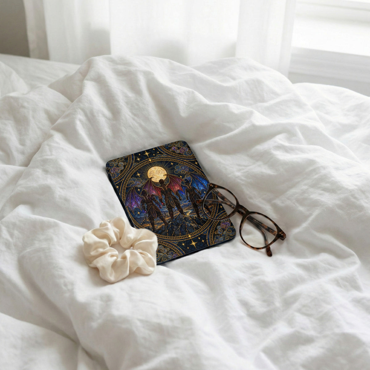 A Kindle case with a fantasy illustration rests on a white bed alongside glasses and a scrunchie.