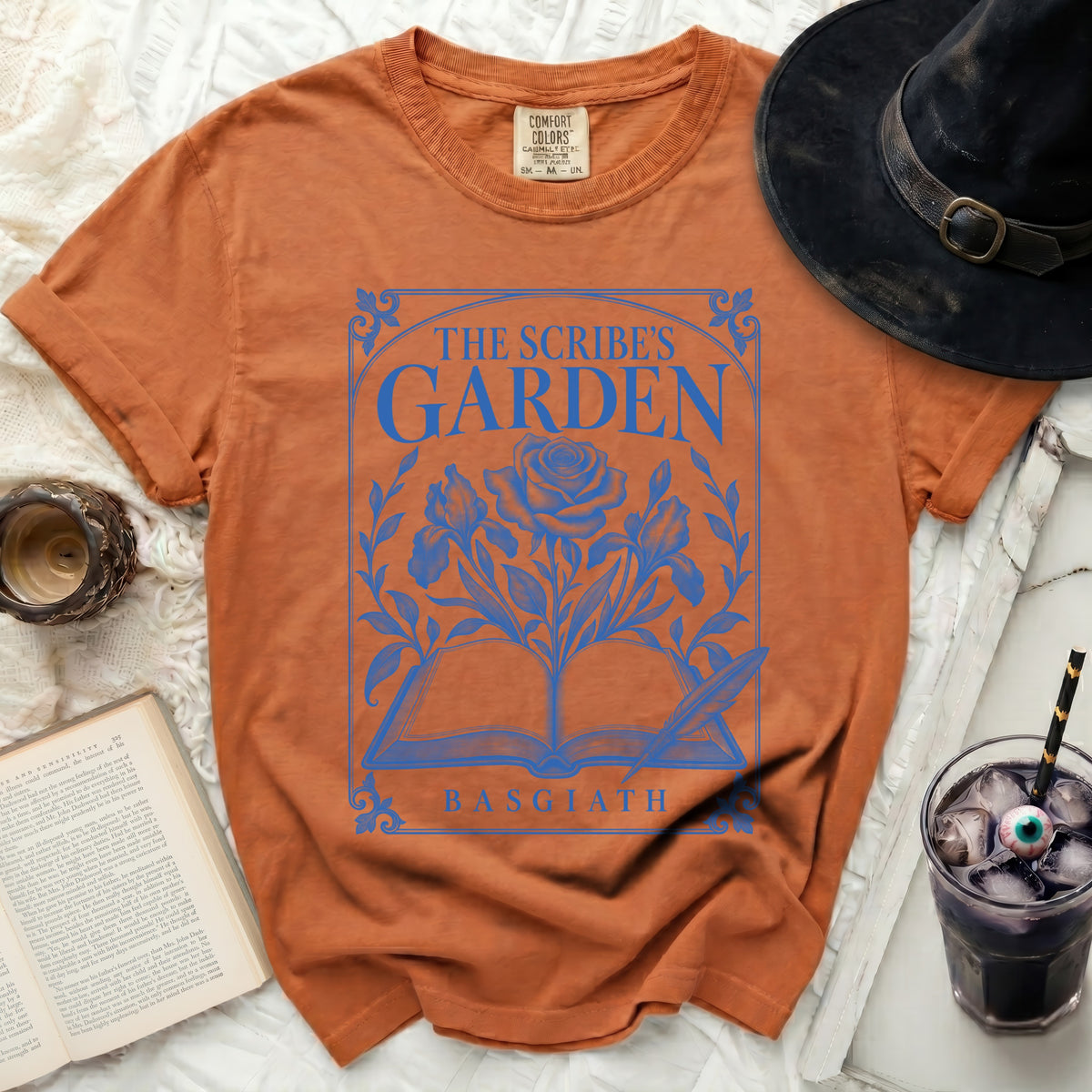 Orange t-shirt with blue graphic of an open book, rose, and text "THE SCRIBE'S GARDEN BASGIATH".
