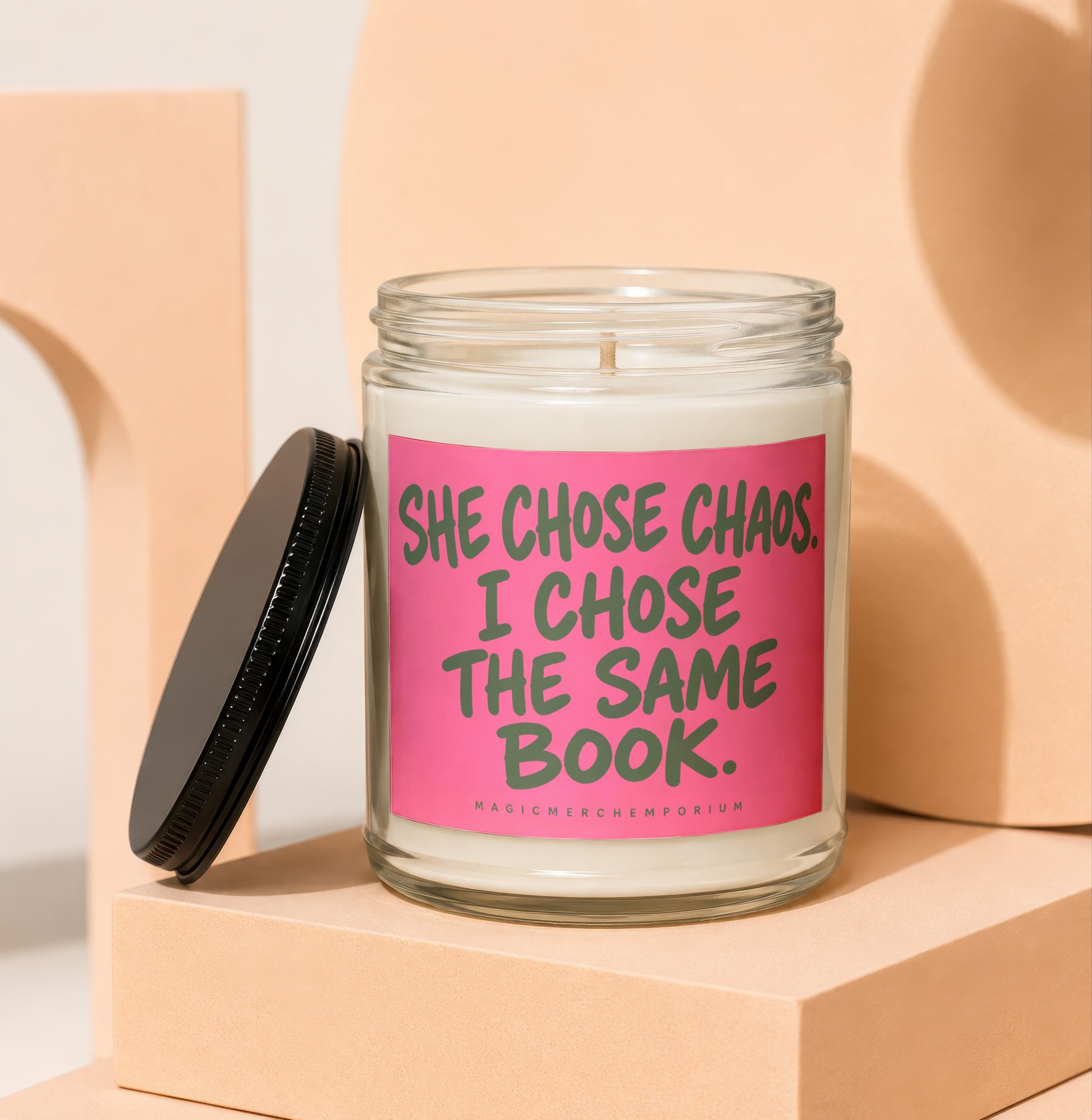 Glass candle with pink label reading "SHE CHOSE CHAOS. I CHOSE THE SAME BOOK."