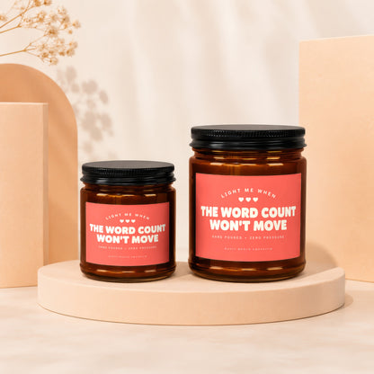 Two amber glass candles with black lids and pink labels that read "The Word Count Won't Move".