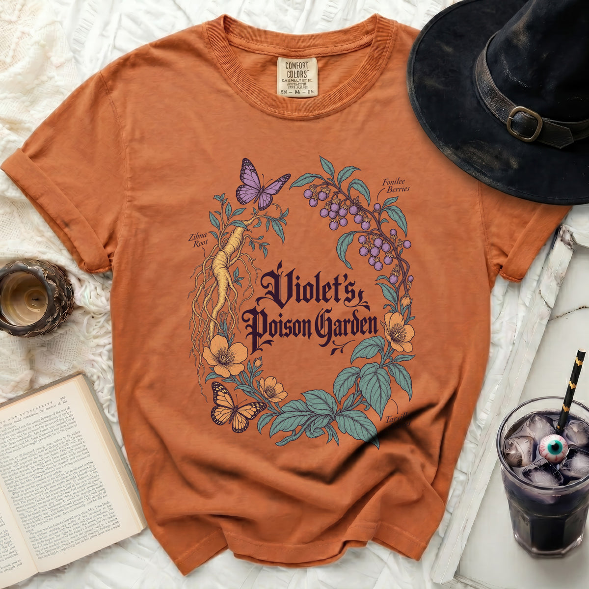 Rust-orange t-shirt featuring a botanical illustration with text reading Violet's Poison Garden, surrounded by a witch hat and book.