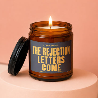 Amber jar candle with black lid, lit flame, text "LIGHT WHEN: THE REJECTION LETTERS COME"