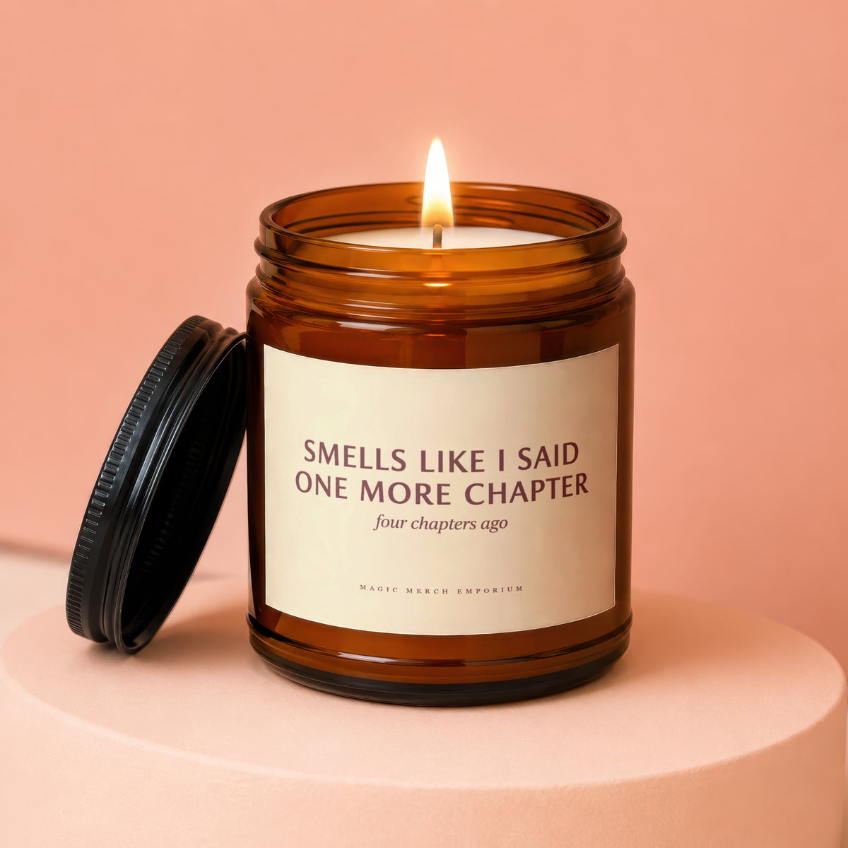 Amber jar candle with a lit wick and lid off, reading "Smells like I said one more chapter".