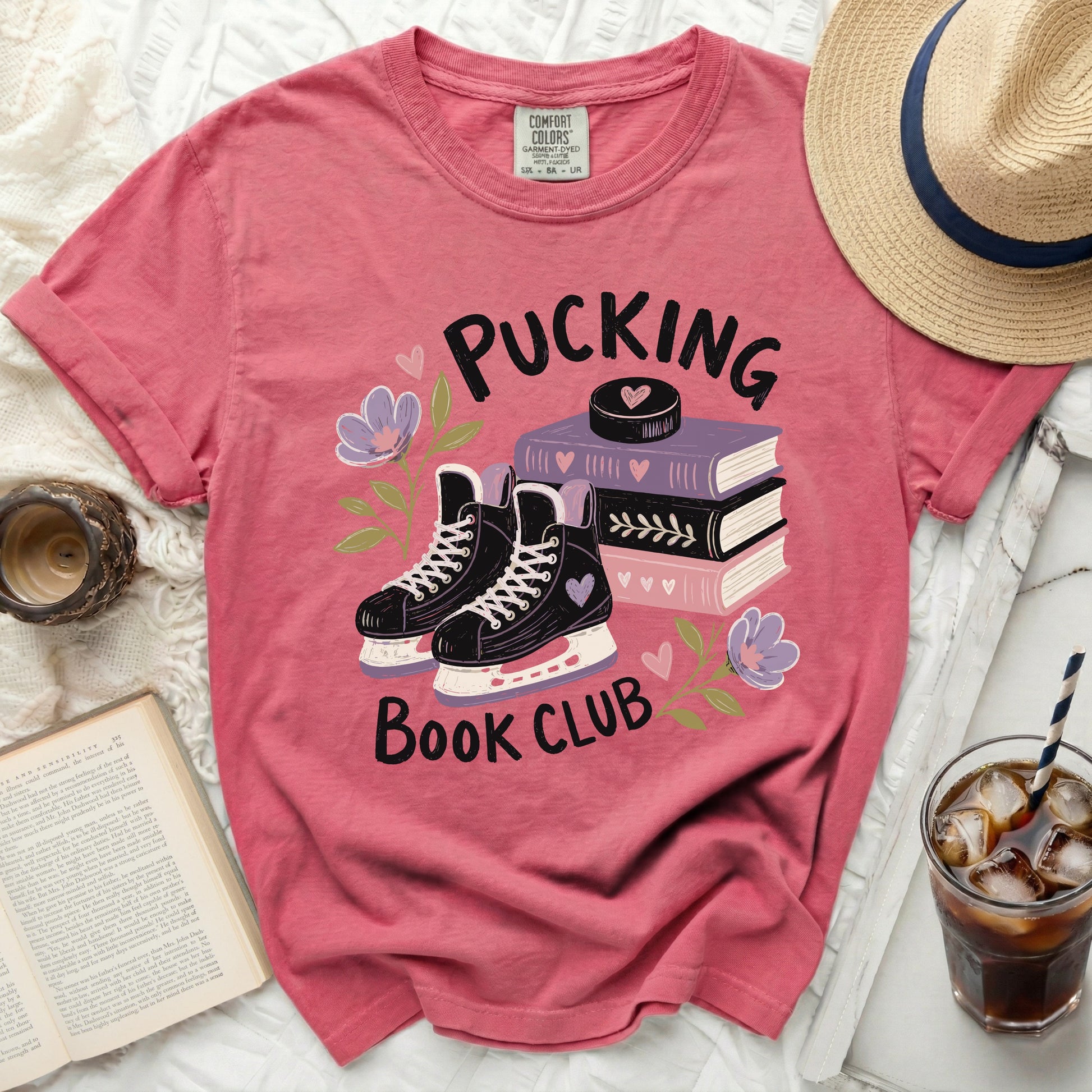 Pink t-shirt with ice skates, books, and flowers, featuring "Pucking Book Club" text.