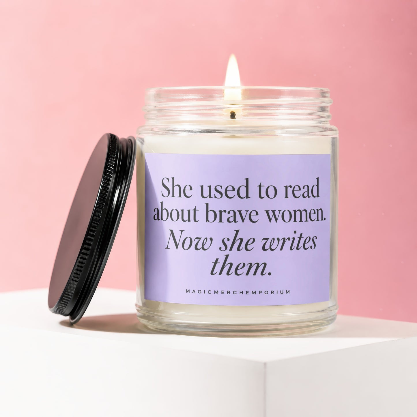 Lit candle in glass jar with purple label, text reads "She used to read about brave women. Now she writes them."