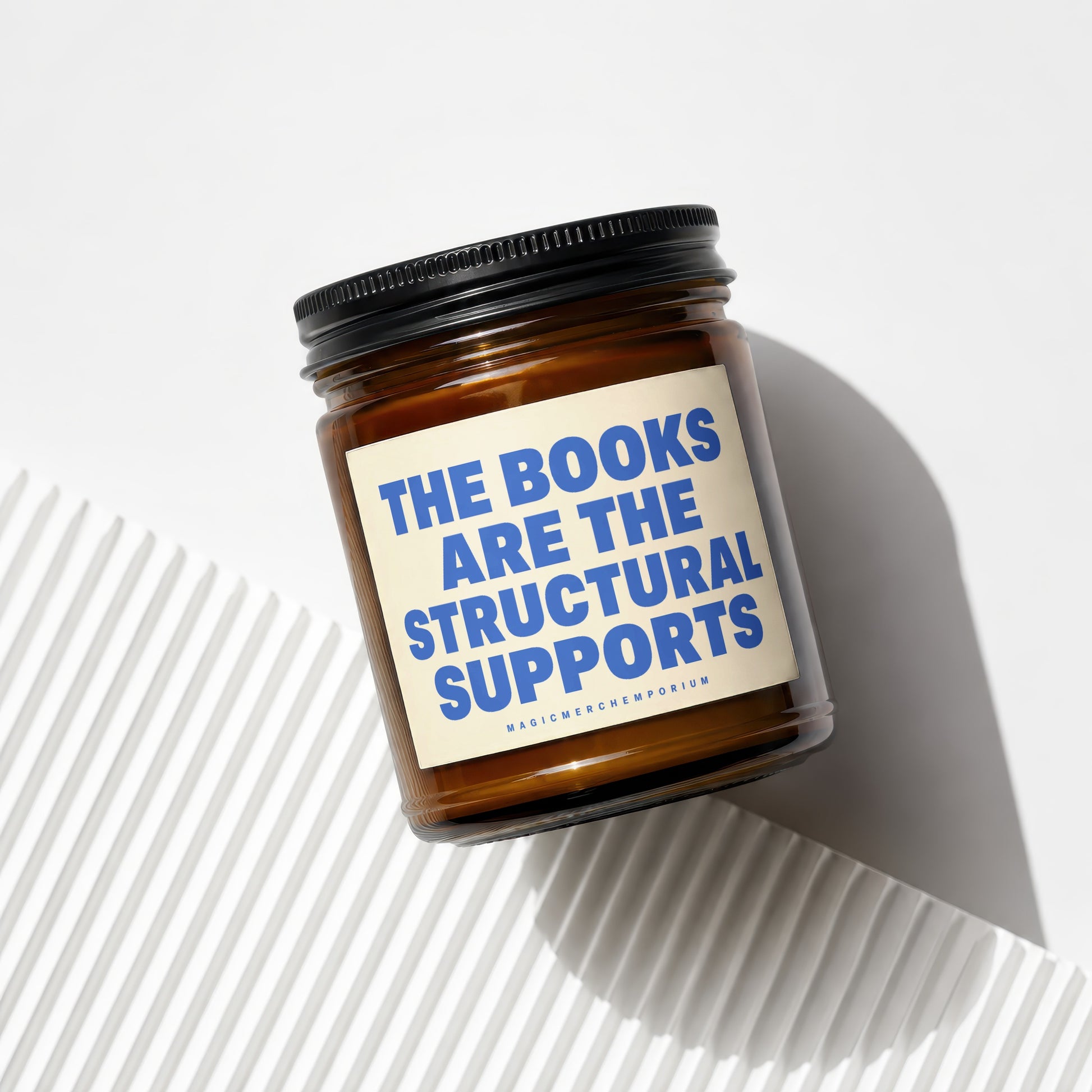 Amber glass candle with black lid featuring text "THE BOOKS ARE THE STRUCTURAL SUPPORTS".