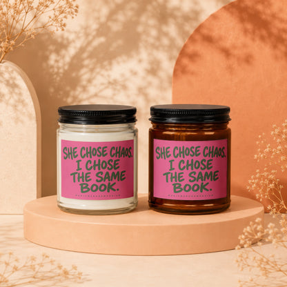 Two candles, one white and one amber, with pink labels reading "SHE CHOSE CHAOS. I CHOSE THE SAME BOOK."