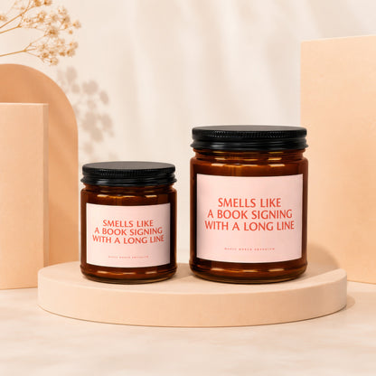Two amber glass candles with pink labels reading "Smells Like A Book Signing With A Long Line".