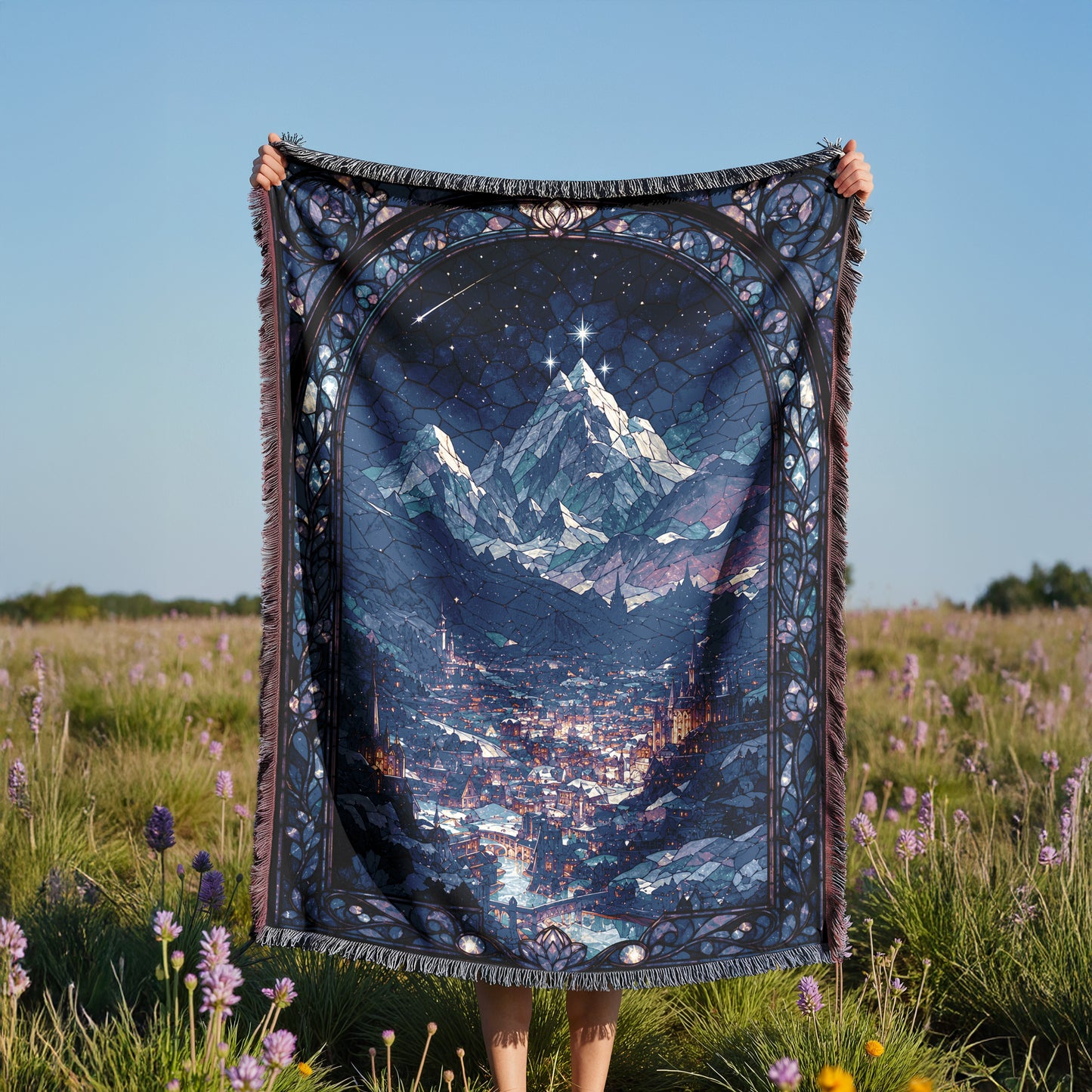 A person is holding up a large, colorful blanket with a mountain landscape printed on it, standing in a field with purple flowers.