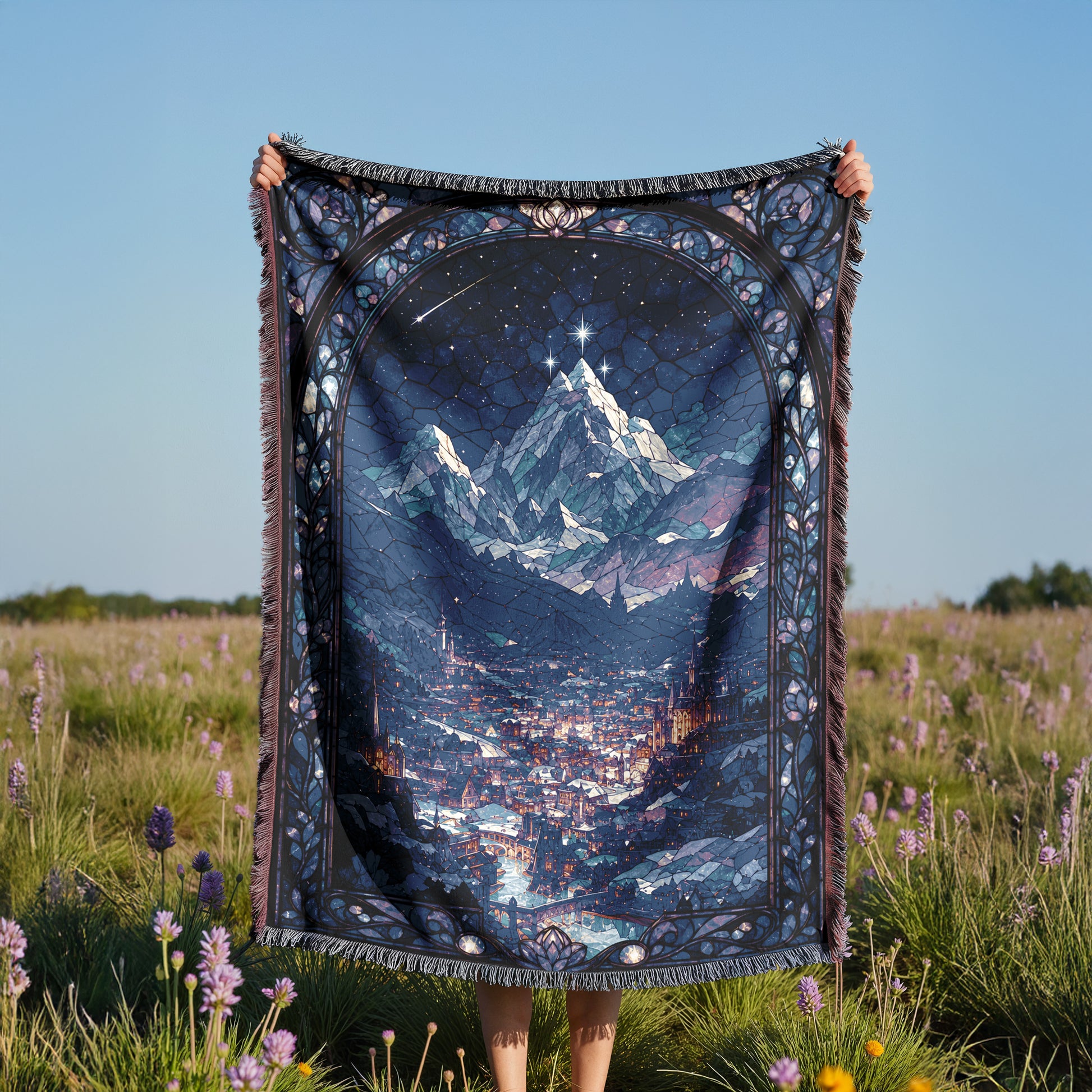 A person is holding up a large, colorful blanket with a mountain landscape printed on it, standing in a field with purple flowers.