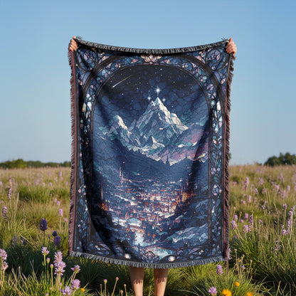 A person is holding up a large, colorful blanket with a mountain landscape printed on it, standing in a field with purple flowers.