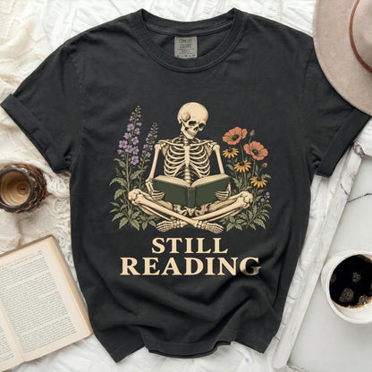 Skeleton reading a book surrounded by flowers with text "Still Reading".