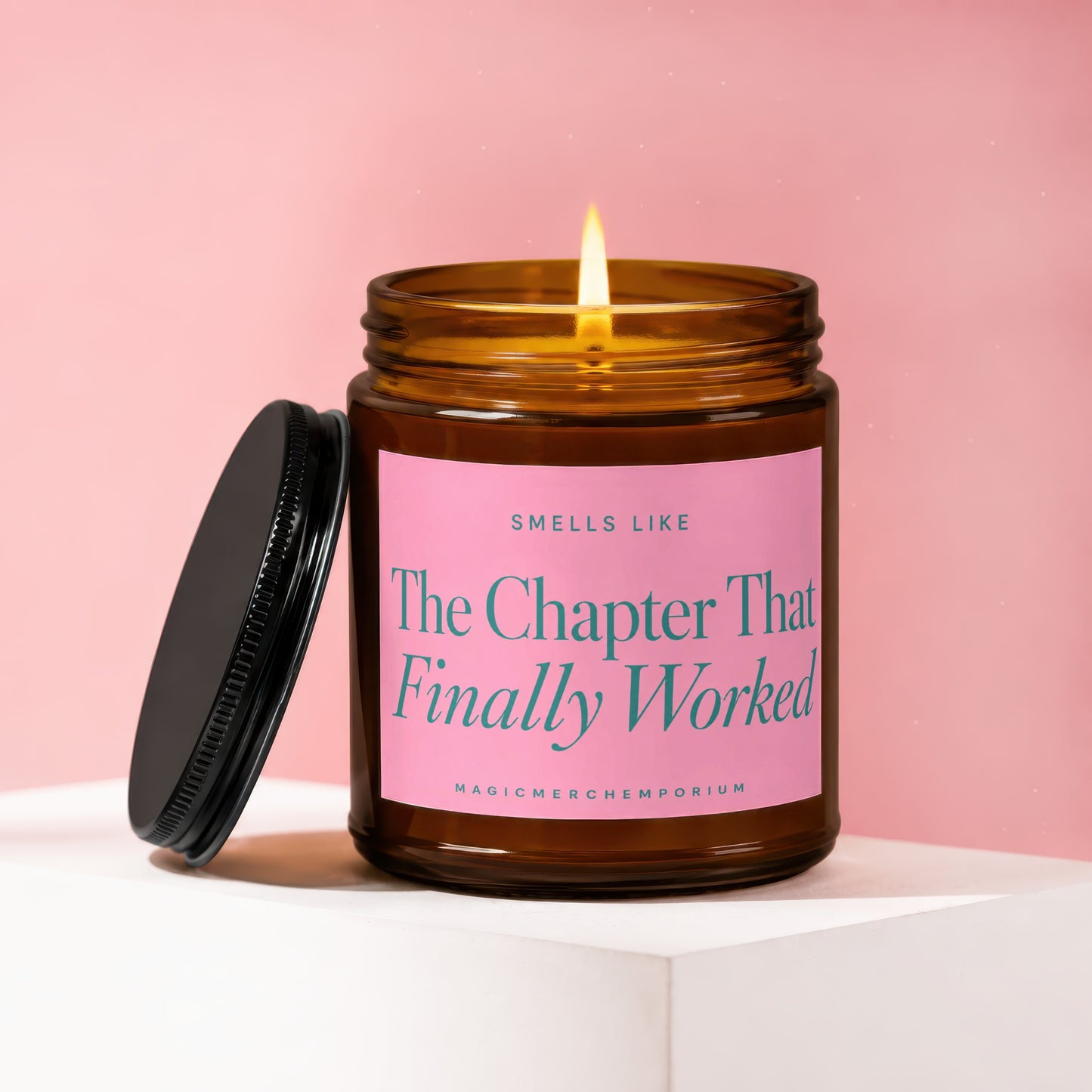 Amber glass candle with lit flame on pink background. Label reads "The Chapter That Finally Worked".
