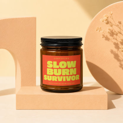 Amber jar candle with a red label reading "CERTIFIED SLOW BURN SURVIVOR MAGICMERCH...EMPORIUM".