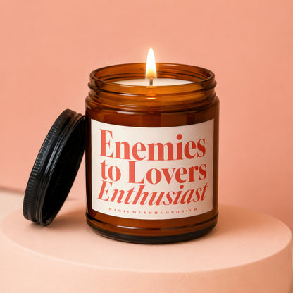 Amber candle in a jar with a lit wick and lid removed, reading "Enemies to Lovers Enthusiast".