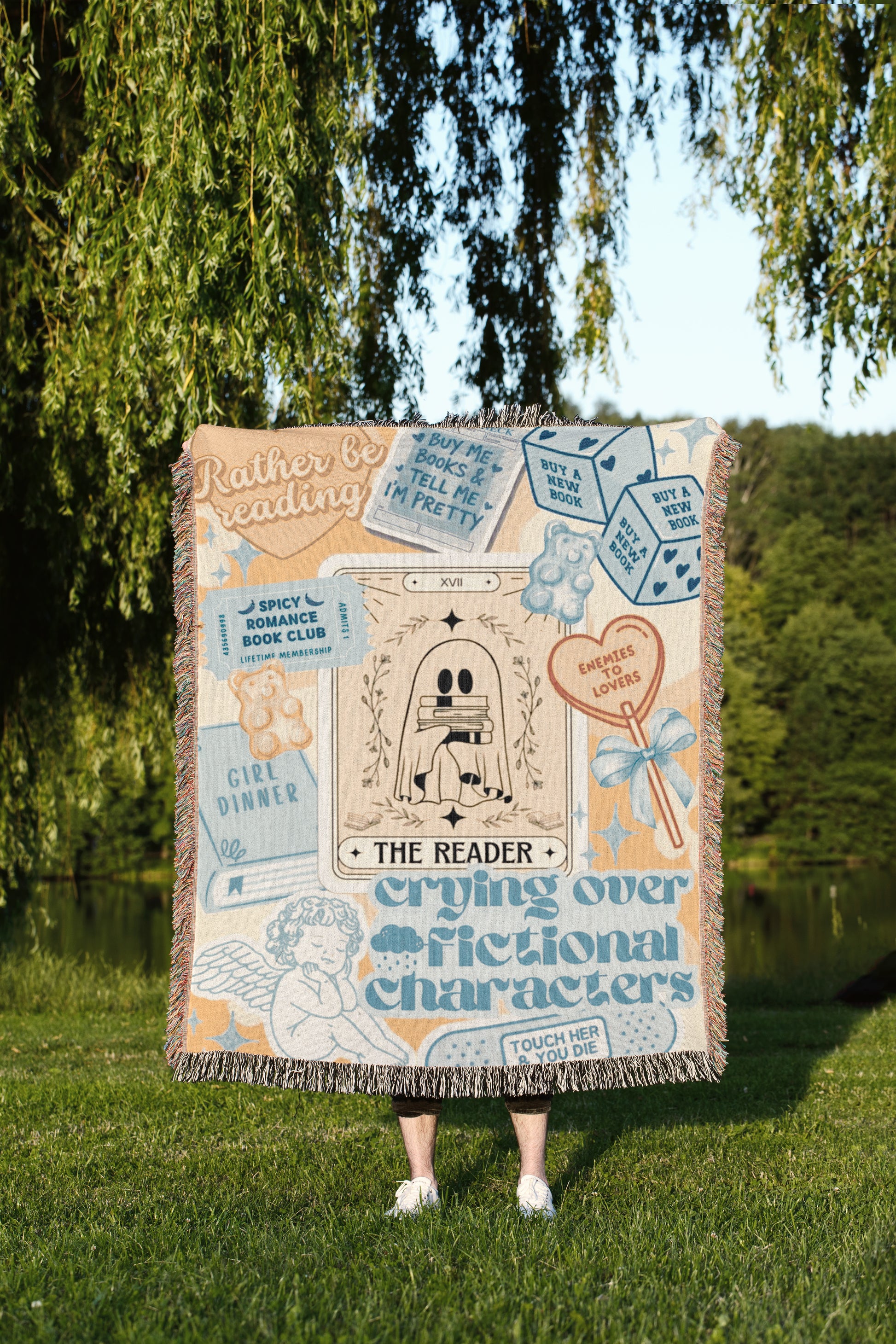 A person is standing in a grassy area, holding a large, colorful blanket that features various text and illustrations, including tarot cards and other decorative elements.