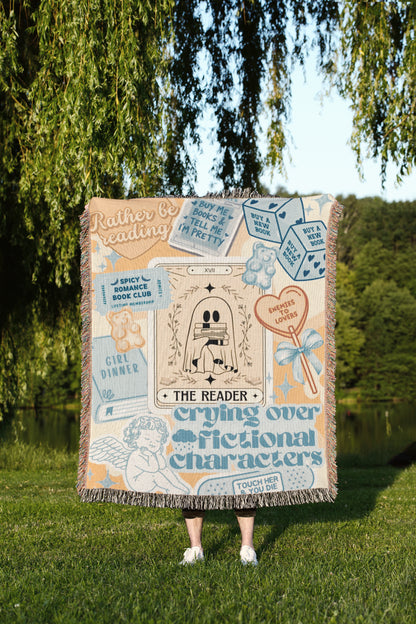 A person is standing in a grassy area, holding a large, colorful blanket that features various text and illustrations, including tarot cards and other decorative elements.