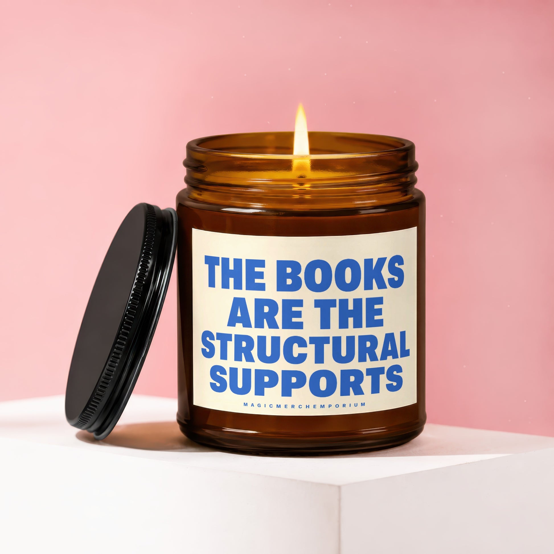 Amber jar candle with lit wick and black lid, reading "THE BOOKS ARE THE STRUCTURAL SUPPORTS."