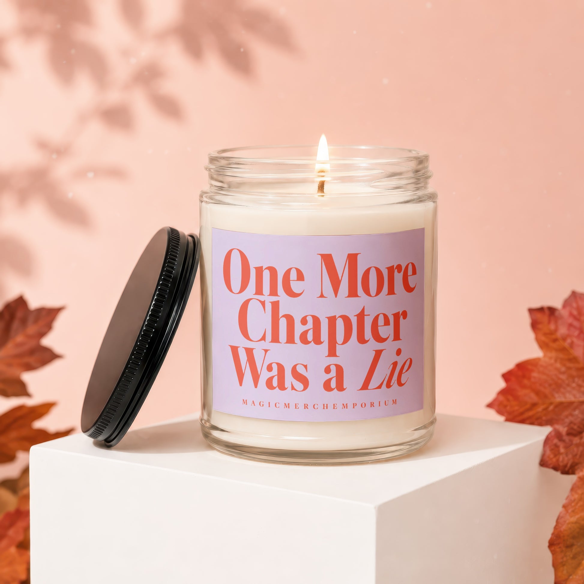 Glass candle with lid off, lit wick, and label reading "One More Chapter Was a Lie".