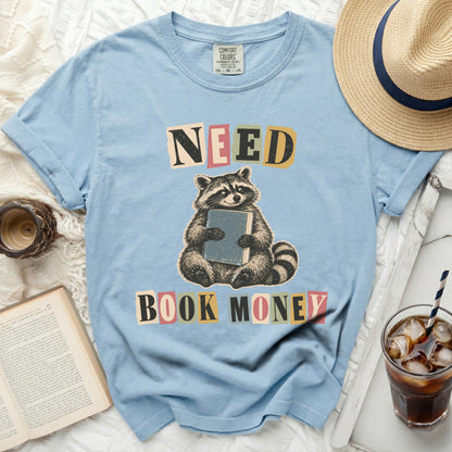 Light blue t-shirt with raccoon holding book and text "NEED BOOK MONEY".