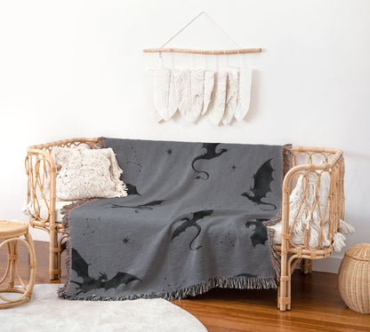 a cozy living room with a gray dragon-themed throw blanket draped over a wicker bench. The room has a white wall with a hanging feather decoration, and a woven basket is placed on the floor.