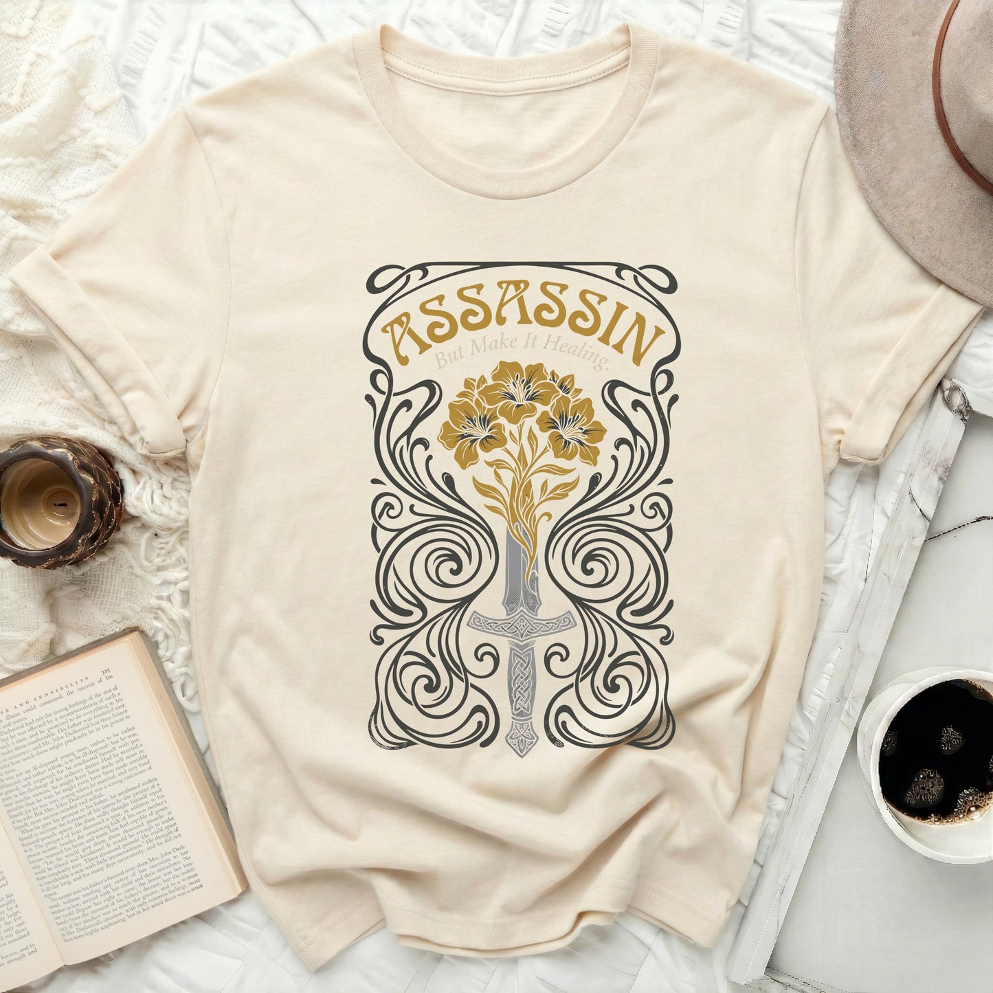 Cream t-shirt with a sword, flowers, and "Assassin But Make It Healing" text.