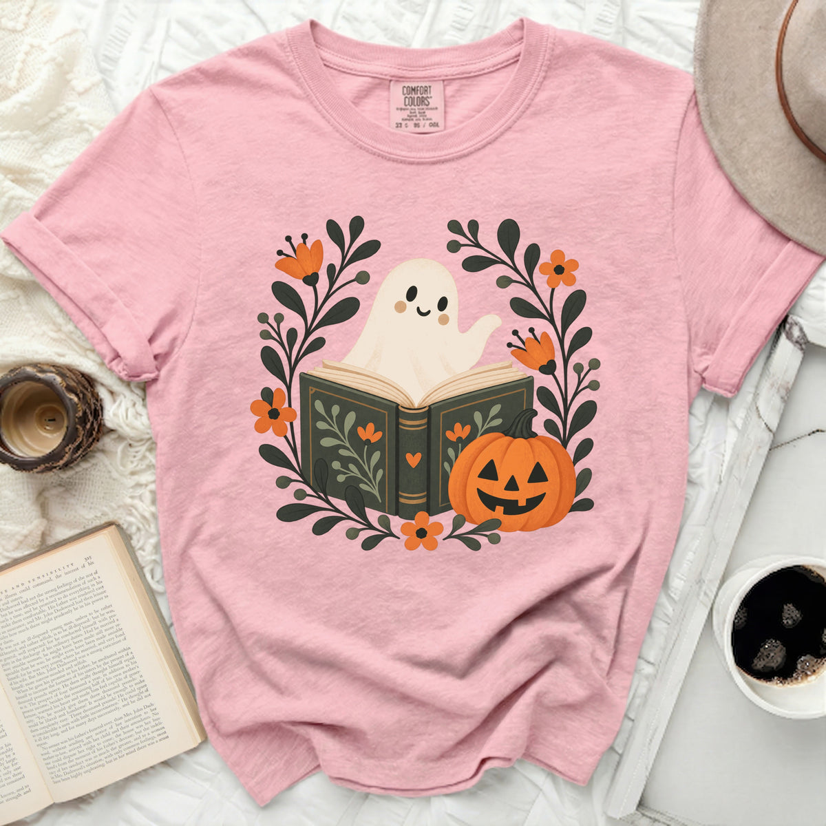 Pink t-shirt with a graphic of a ghost reading a book surrounded by pumpkins and flowers.
