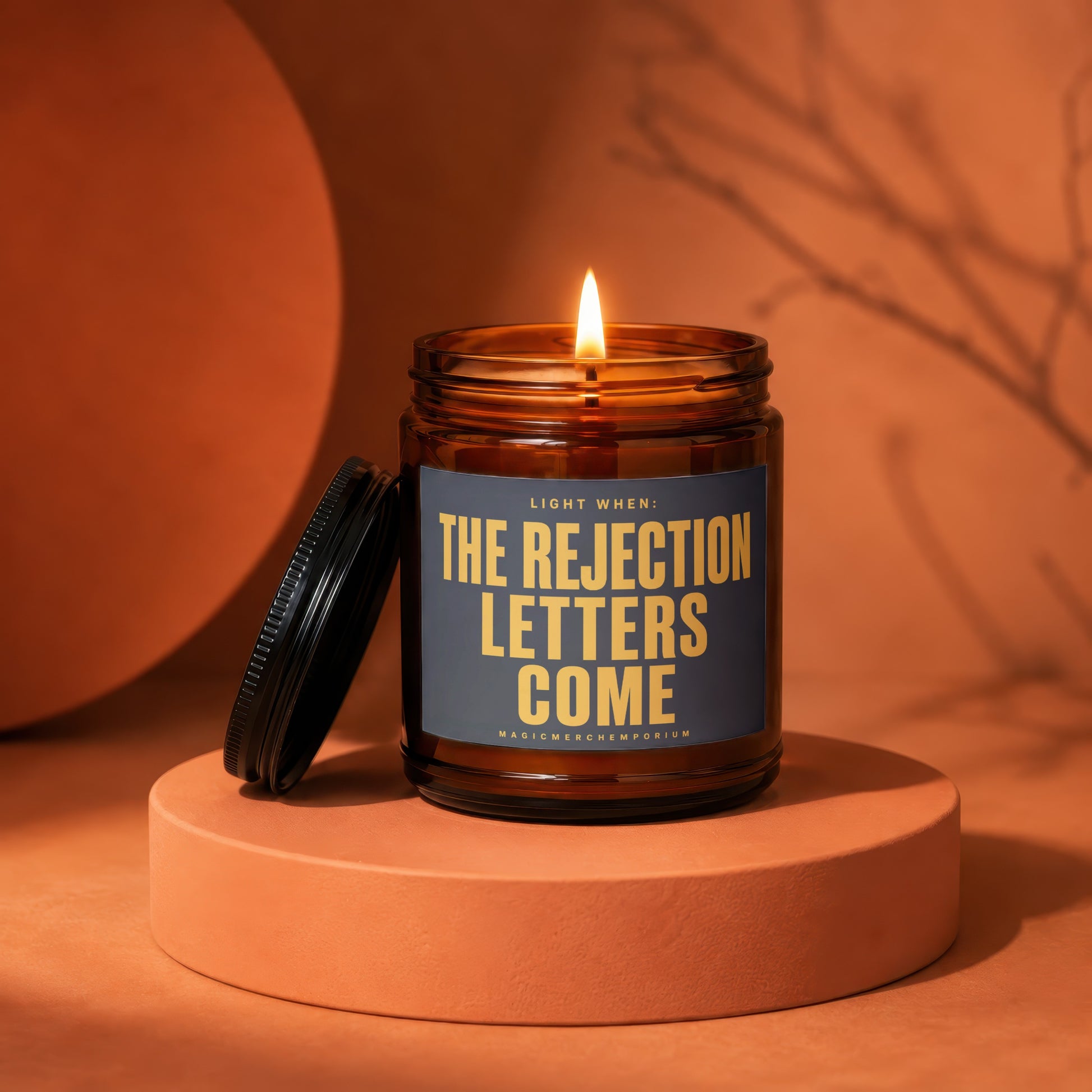 Amber jar candle with lid off, burning and displaying text "Light When: The Rejection Letters Come".