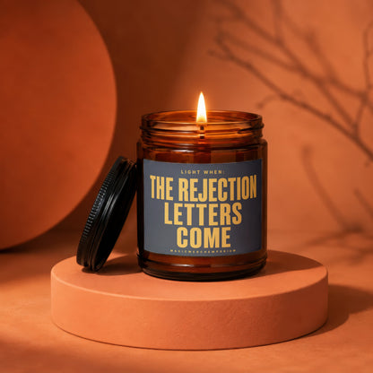 Amber jar candle with lid off, burning and displaying text "Light When: The Rejection Letters Come".