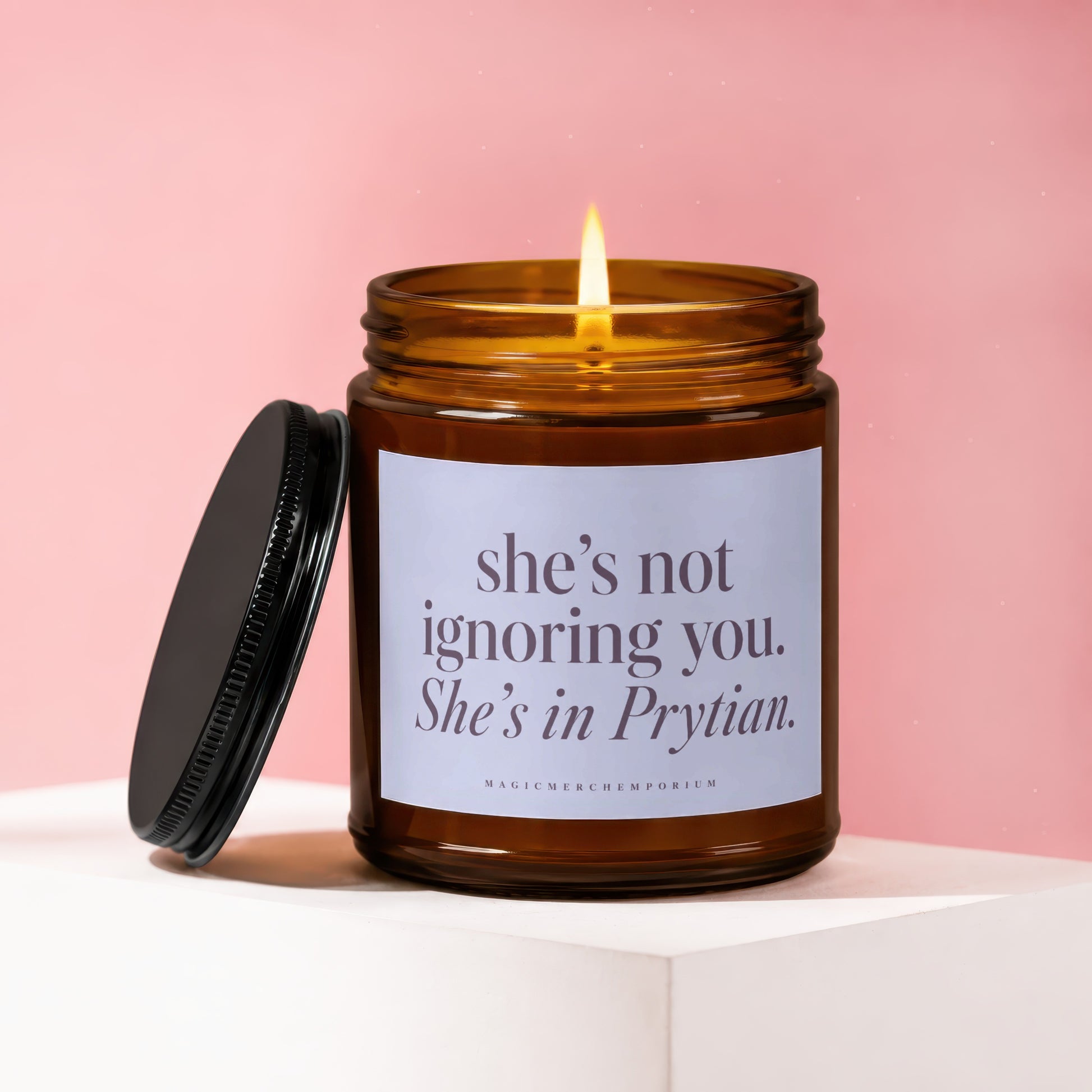 A lit brown glass jar candle with a black lid resting next to it, featuring a label that reads "She's not ignoring you. She's in Prytian."