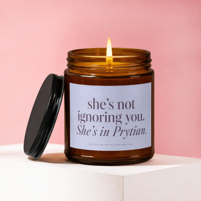 A lit brown glass jar candle with a black lid resting next to it, featuring a label that reads "She's not ignoring you. She's in Prytian."