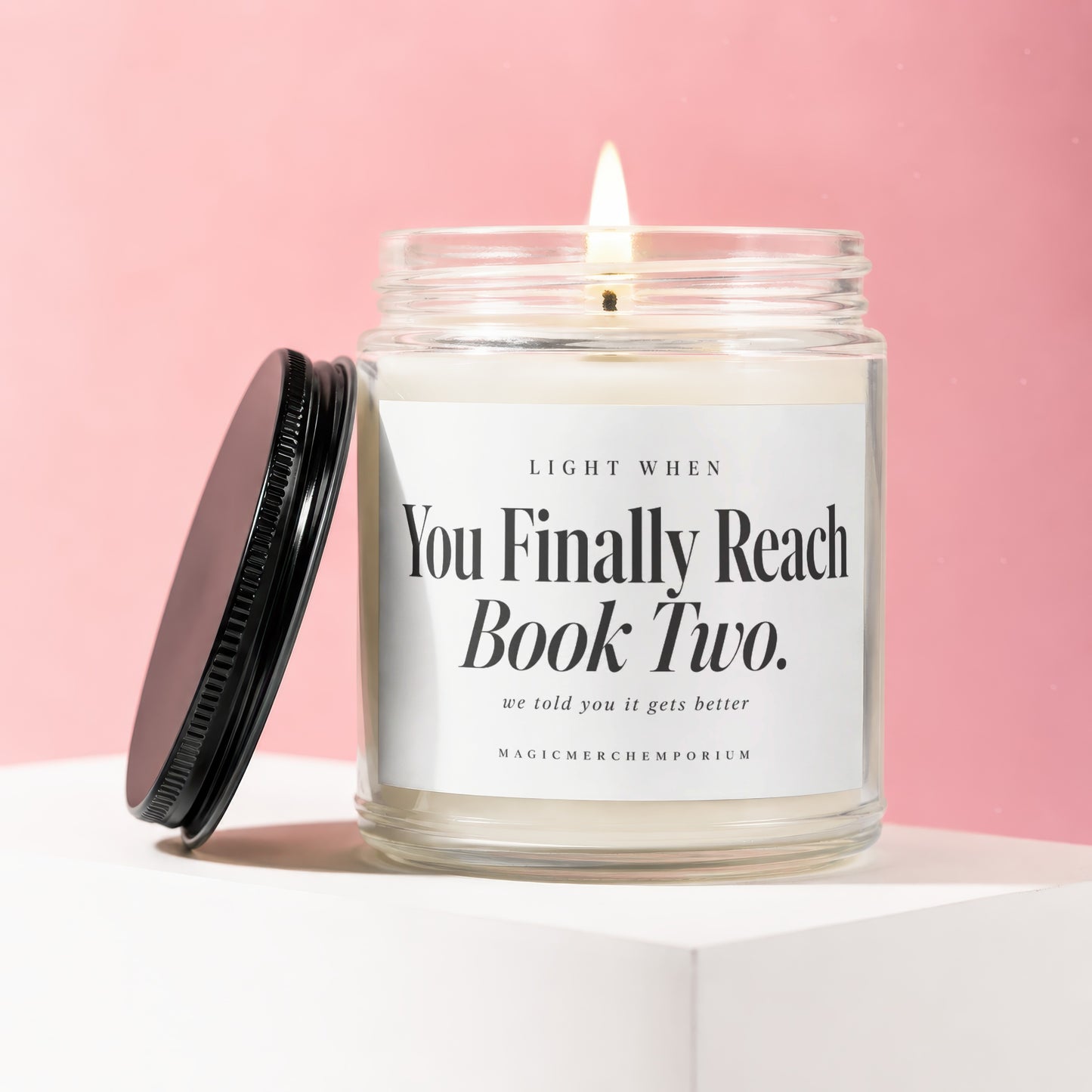 A lit soy candle in a glass jar with a black lid and white label against a pink background.
