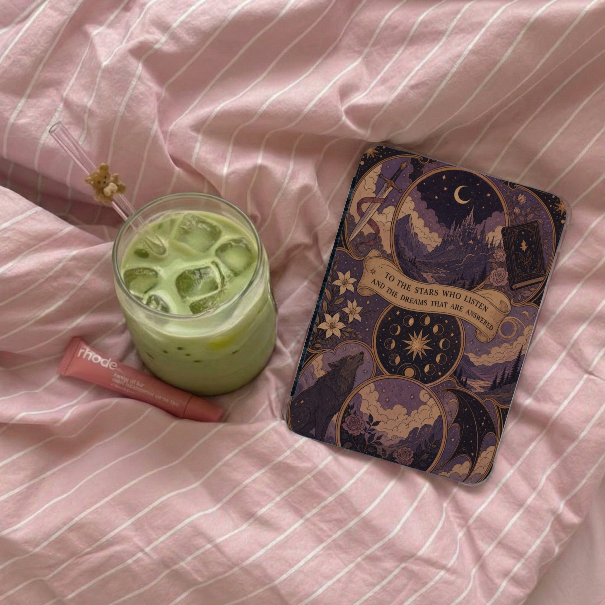 Phone case with celestial, wolf, and castle design next to iced matcha on pink striped bedding.