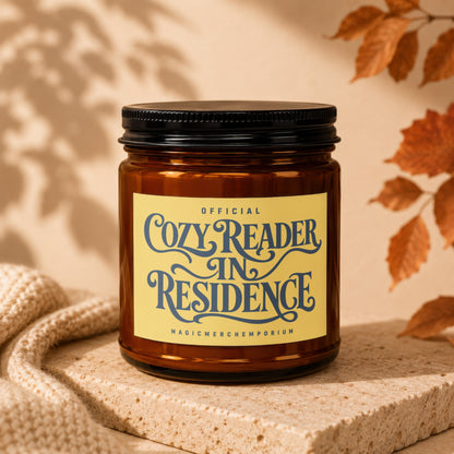 Amber jar candle with a yellow label that reads "Official Cozy Reader In Residence MagicMerchEmporium."