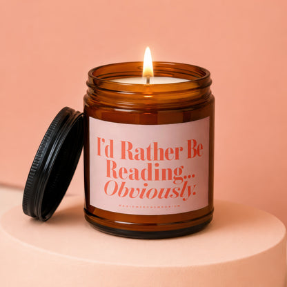 Amber jar candle with lit flame and lid beside it, reads "I'd Rather Be Reading... Obviously".