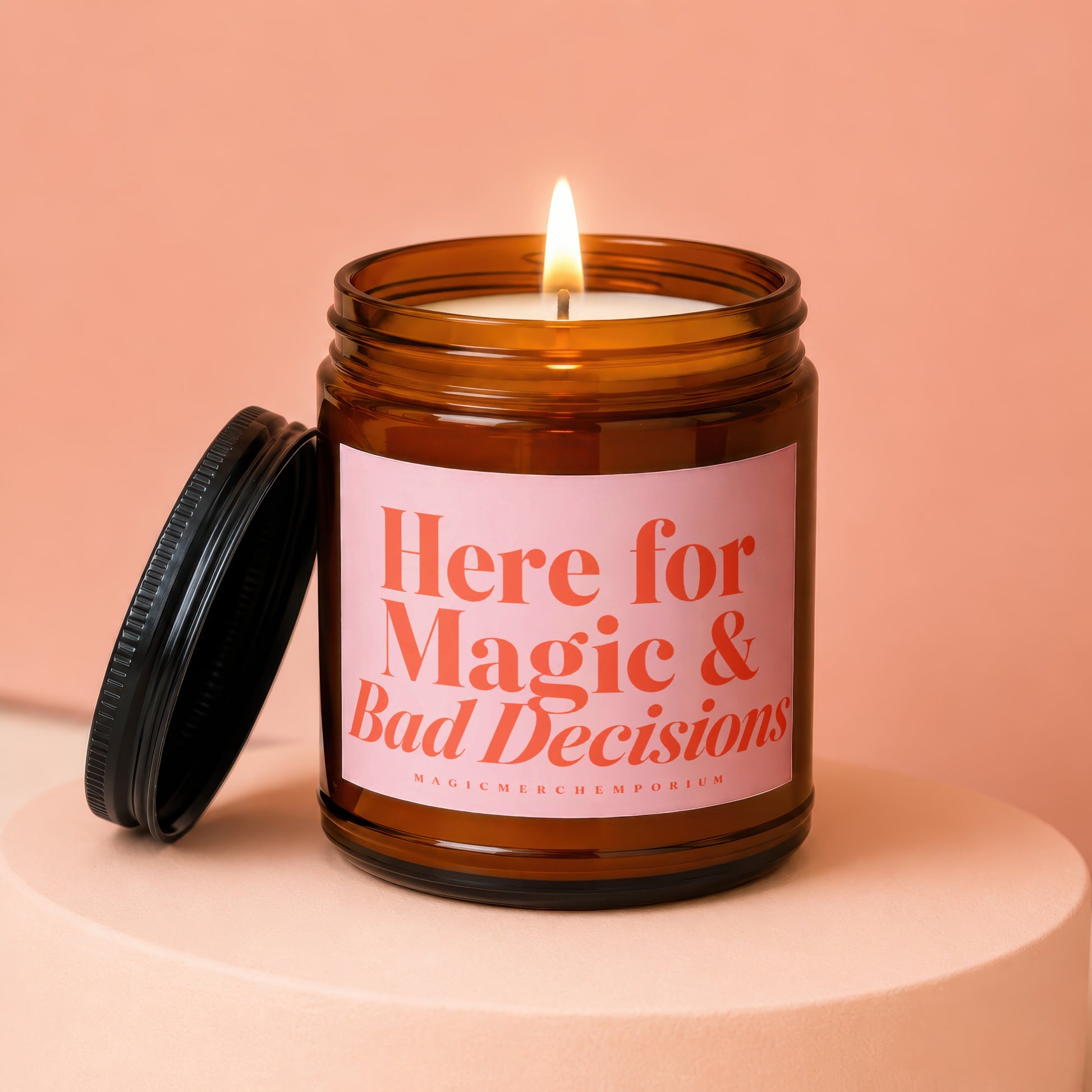 Amber jar candle with lit wick and pink label reading "Here for Magic & Bad Decisions".